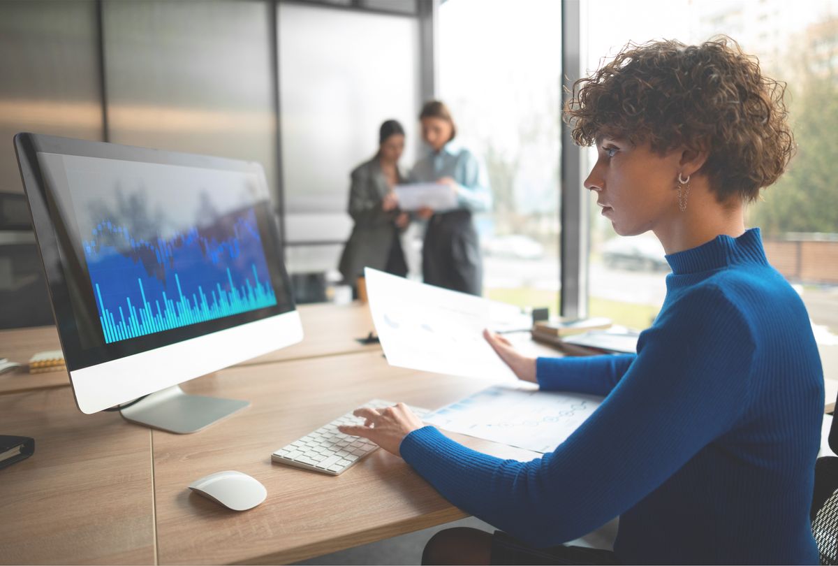 Una mujer está trabajando en su computadora en una oficina con grandes ventanas mientras dos personas conversan al fondo.