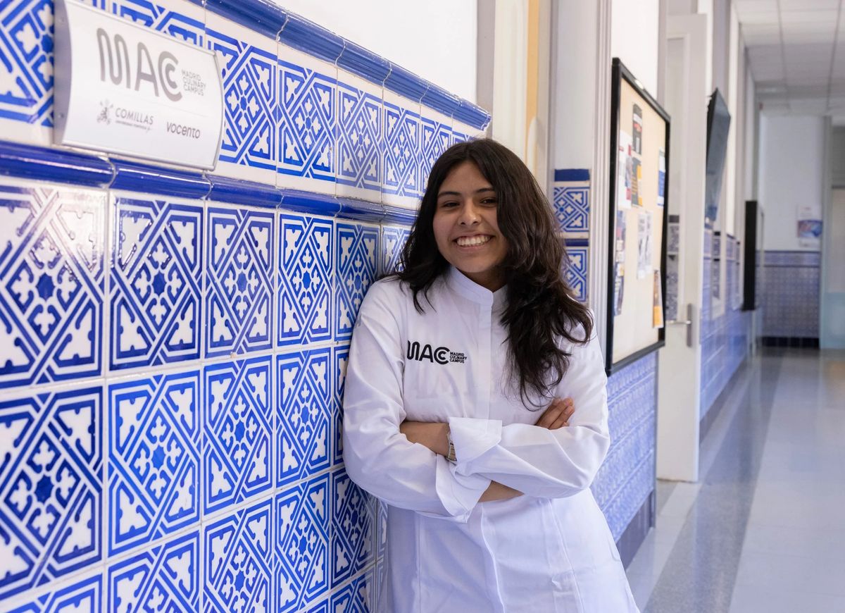 Una joven sonriente con uniforme blanco se apoya en una pared decorada con azulejos azules en un pasillo escolar.