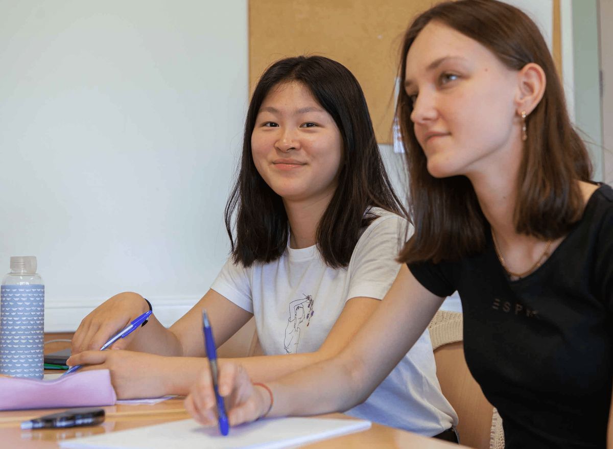 Dos jóvenes sentadas en un aula, sonriendo mientras escriben en sus cuadernos.