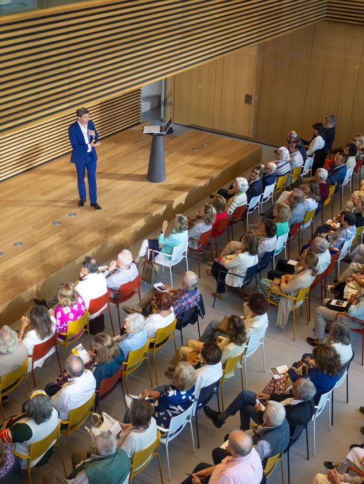 Un orador presenta frente a un gran público en un auditorio moderno con asientos coloridos.