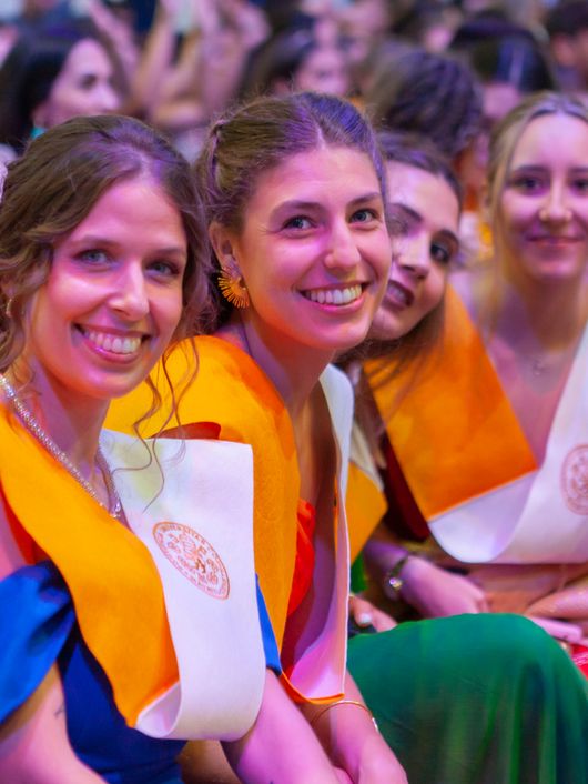 A group of young graduates wearing colorful stoles smiling at a graduation ceremony.