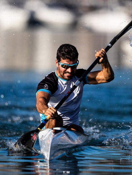 Un hombre compite en piragüismo a velocidad en aguas tranquilas, remando intensamente con un fondo de barcos desenfocados.