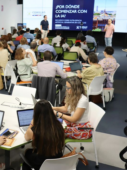 A group of adults are sitting around tables in a conference room setting, engaged in discussion and looking at laptops, with a presentation screen in the background.