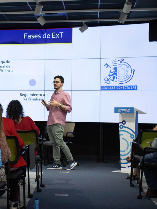 A man presents at a workshop in a conference room with attendees watching and a digital screen displaying a presentation.