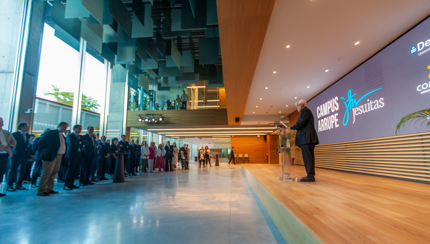 A speaker presenting in front of an audience at a modern indoor venue with digital signage displaying the text 'Campus Arrupe Jesuitas'.