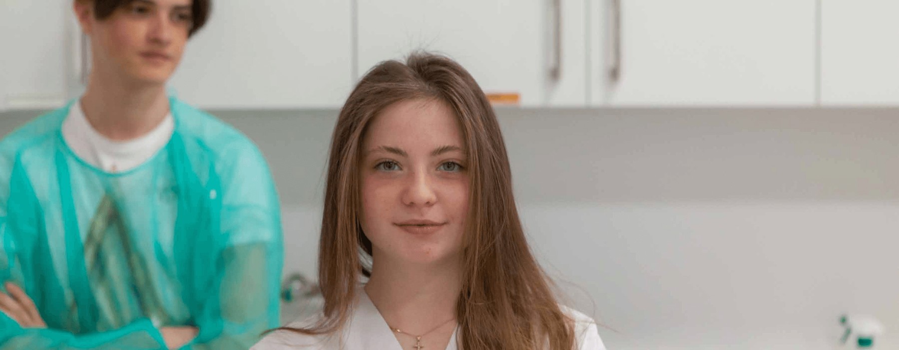 A young woman in a white lab coat standing confidently in a lab setting with a male colleague in green scrubs in the background.