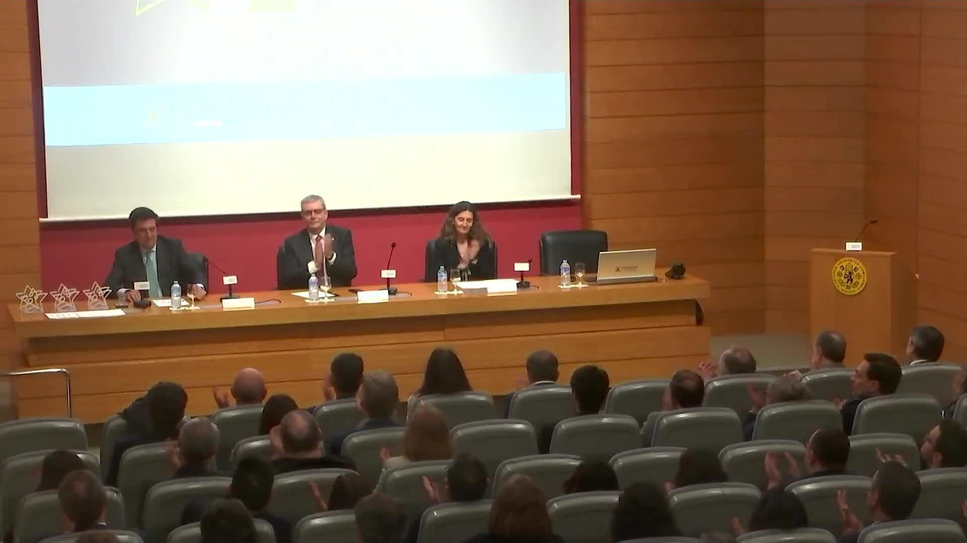 Una conferencia con tres ponentes en un podio y un público aplaudiendo.