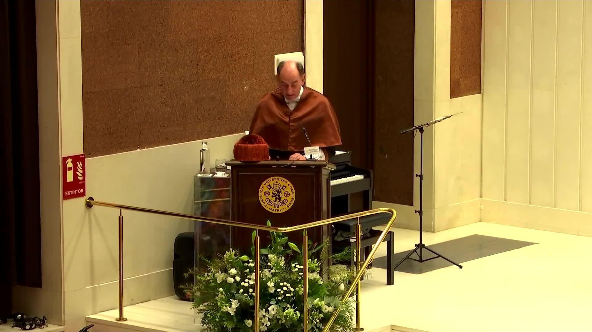 A man in a brown robe speaking at a podium inside a room with a floral arrangement in front.