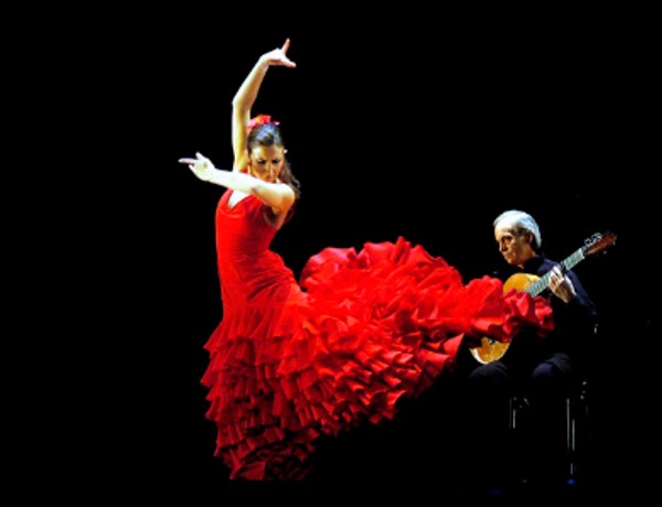 Una bailarina de flamenco con un vestido rojo vibrante realiza una pose enérgica mientras un guitarrista la acompaña.