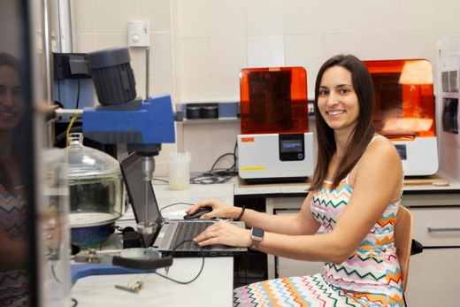 A woman smiling while working on a laptop in a laboratory with 3D printers in the background.