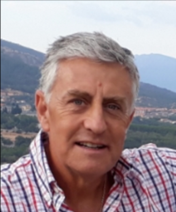 Retrato de un hombre mayor con cabello canoso y camisa a cuadros, sonriendo frente a un fondo de montañas.
