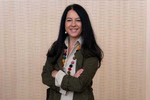 A woman in a green jacket and colorful necklace is standing with crossed arms, smiling at the camera against a textured beige background.