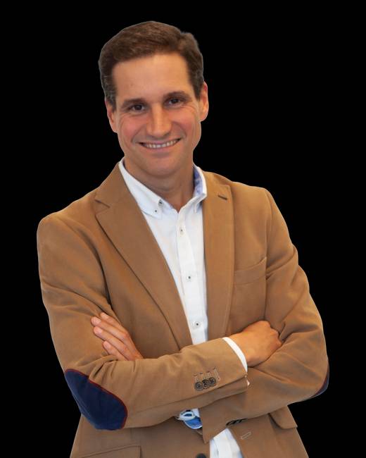 Un hombre sonriente con una chaqueta marrón y una camisa blanca, posando con los brazos cruzados.