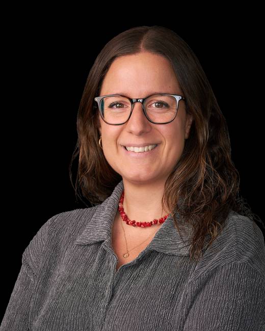 Retrato de una mujer sonriente con gafas y cabello ondulado, vestida con una camisa gris.