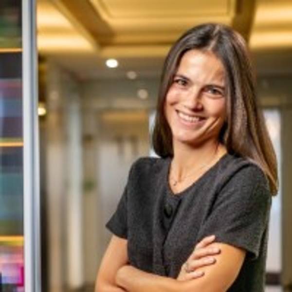Una mujer sonriente con cabello largo y oscuro, vestida con una blusa negra, posa con los brazos cruzados en un entorno moderno.