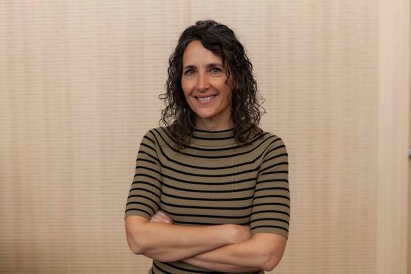 Una mujer sonriente con cabello rizado usa una camiseta de rayas y está de pie con los brazos cruzados frente a un fondo neutro.