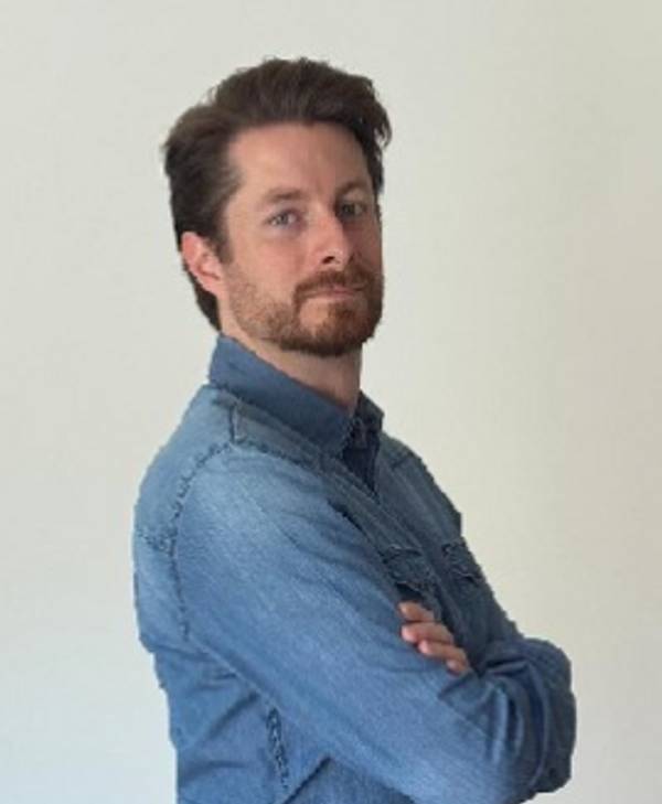 Un hombre con camisa de mezclilla y barba posando con los brazos cruzados.