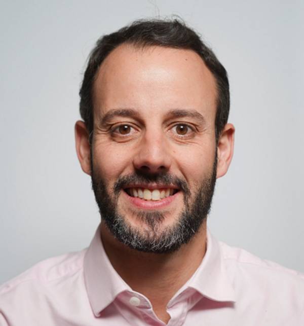 Un hombre sonriente con barba de manera casual frente a un fondo claro.
