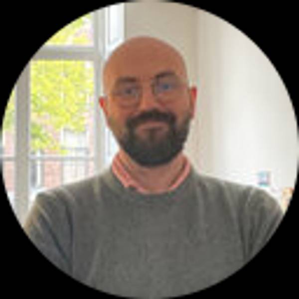 Retrato de un hombre con barba y gafas sonriendo en un entorno luminoso.