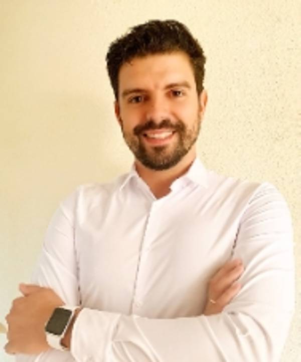 A smiling man with dark hair and a beard, wearing a white shirt and crossing his arms.