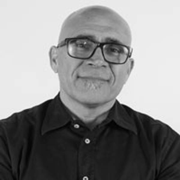 Un hombre con gafas y barba, vestido con una camisa negra, sonríe frente a un fondo blanco.