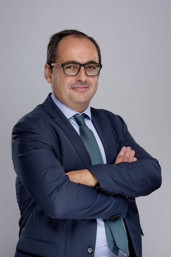 Un hombre vestido formalmente con gafas y una corbata, posando con los brazos cruzados.