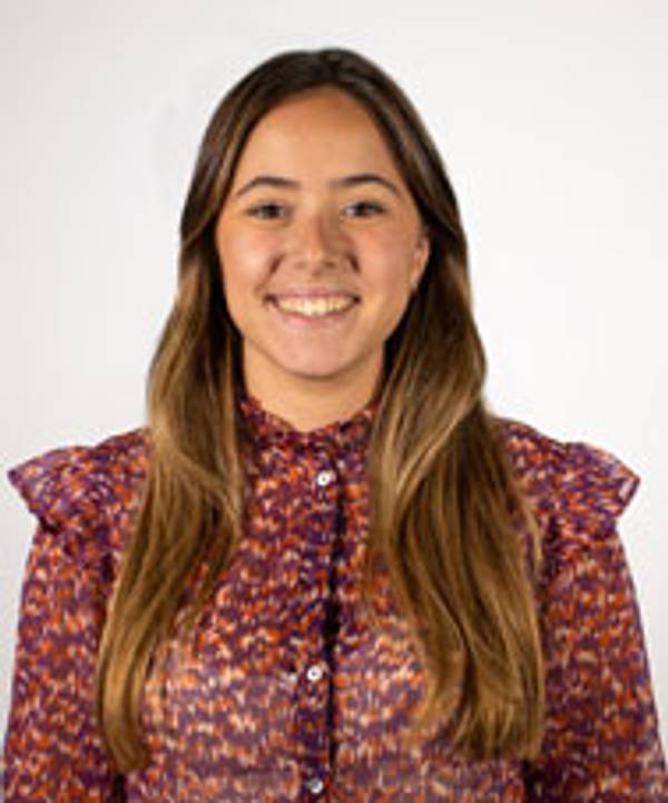 A young woman smiling at the camera wearing a patterned blouse against a white background.