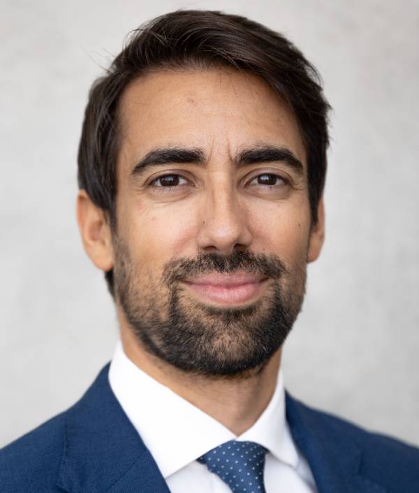 Retrato de un hombre sonriente con barba, vestido con un traje azul y corbata.
