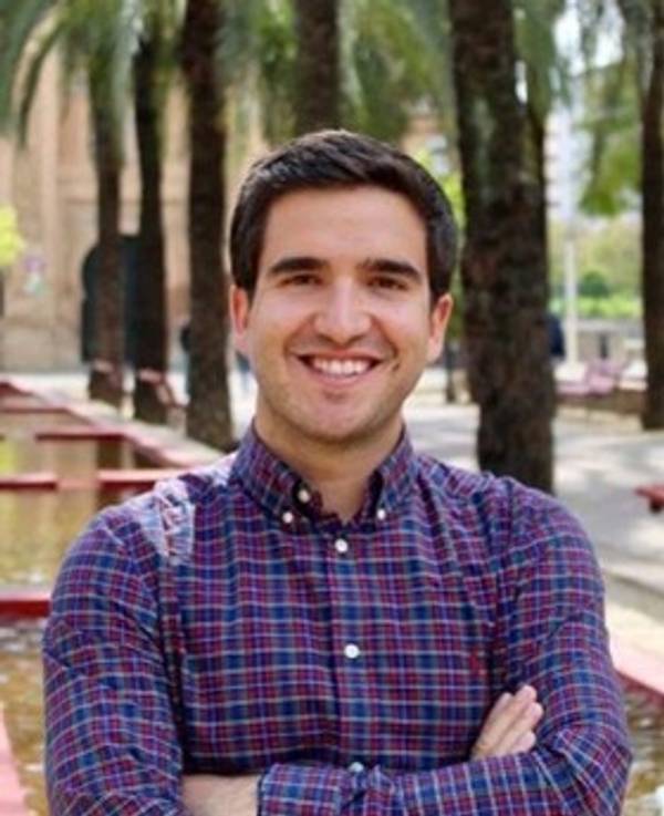 Un hombre sonriente con camisa a cuadros posa en un parque con palmeras de fondo.