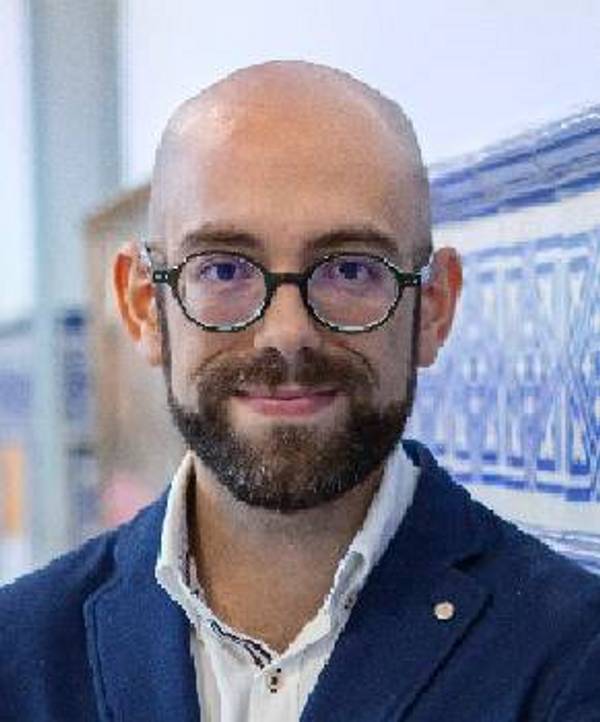 A bald man with a beard wearing glasses and a blue jacket smiles in front of an indistinct background.