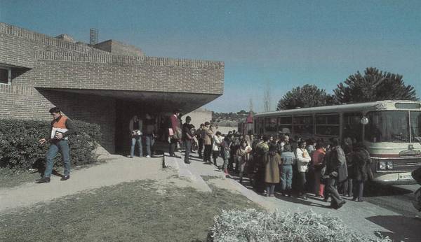 A group of people boarding a bus outside a modern building under a clear sky.