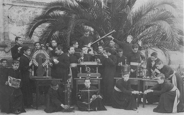 Historical photo of a group of priests with various scientific instruments, some engaged in observation and others in discussion, set against an outdoor backdrop with palms.