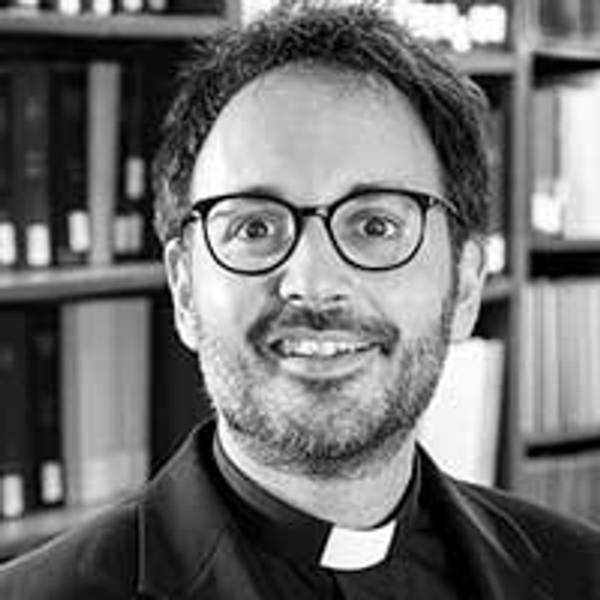 Retrato en blanco y negro de un hombre con gafas y un collar de clérigo sonriendo frente a una estantería de libros.
