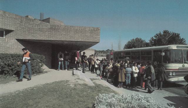 Personas subiendo a un autobús frente a un edificio de ladrillo con arquitectura característica.