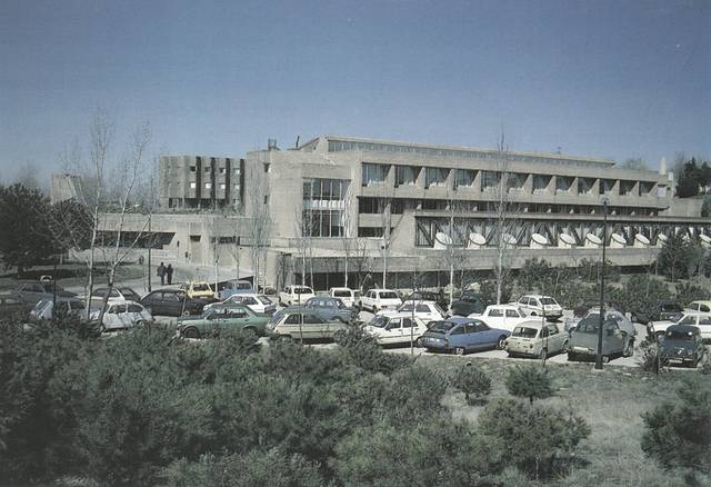 Fotografía de un edificio de varios pisos con un estacionamiento lleno de coches en primer plano.