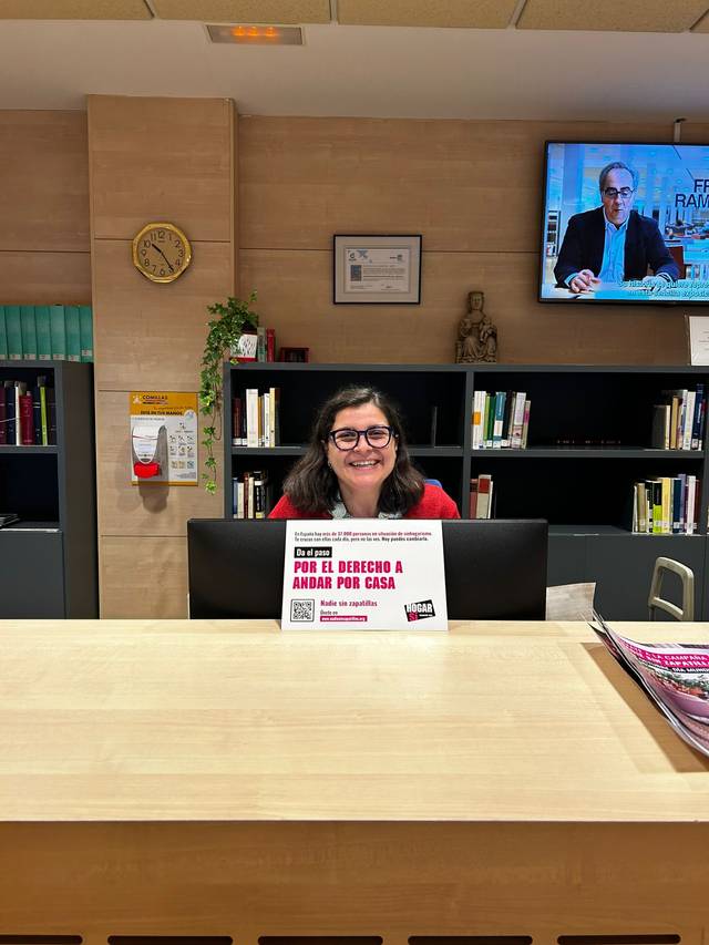 A woman smiling at a reception desk with a bookshelf and a television screen in the background.