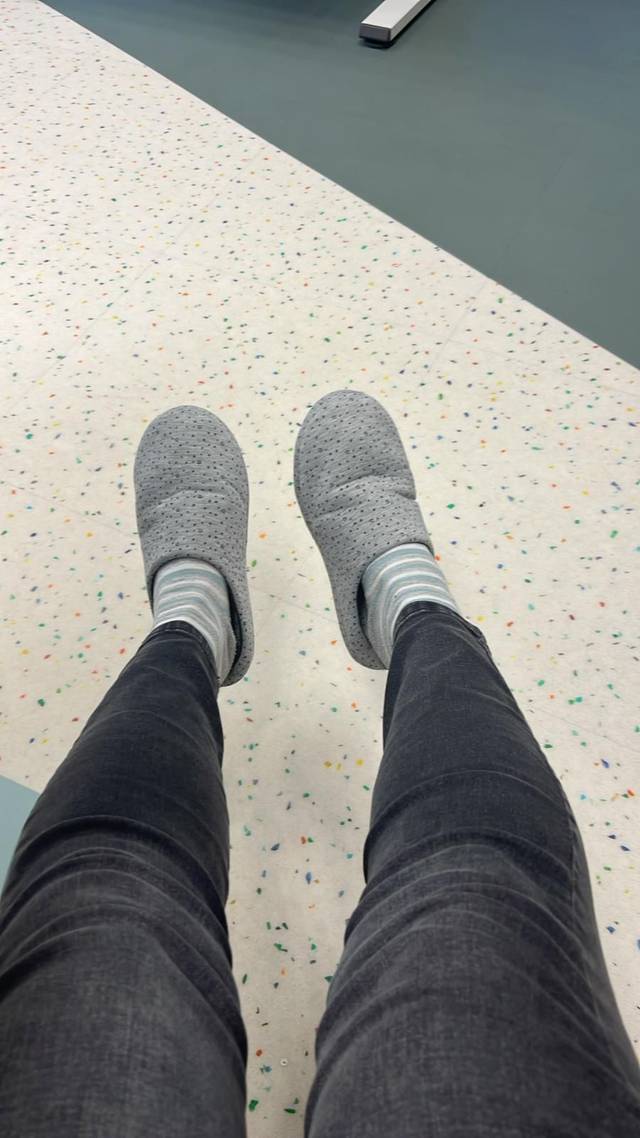 A person wearing gray slip-on shoes and jeans, sitting with their feet facing forward on a speckled floor.