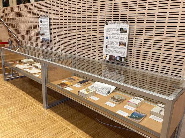 Una exposición de documentos y libros dispuestos en una vitrina de vidrio con una pared perforada de madera al fondo.