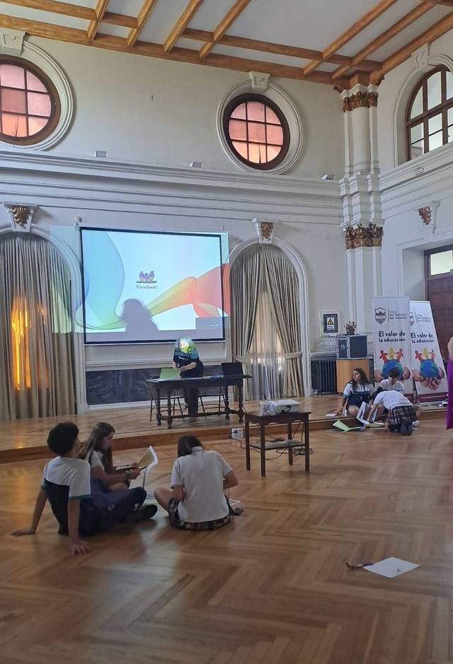 Un grupo de niños y adolescentes participan en una actividad en un salón con un proyector y mobiliario sencillo.