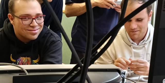 Dos jóvenes concentrados en una actividad frente a un ordenador, rodeados de cables y pantallas.