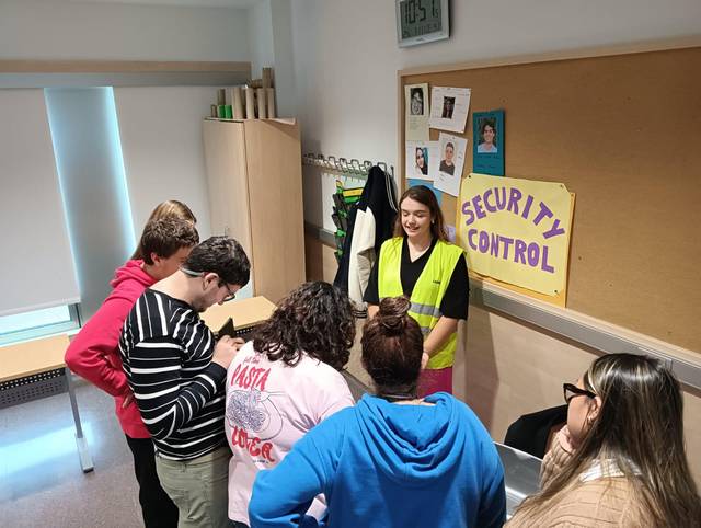 Un grupo de personas interactúa con una mujer que lleva un chaleco de seguridad en un ambiente interior.