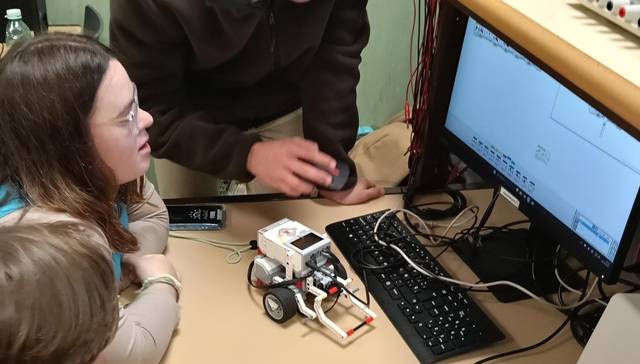 Un grupo de personas observa un robot mientras interactúan con una computadora en un entorno educativo.