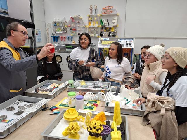 Un grupo de personas observa con interés una demostración sobre impresoras 3D en un entorno educativo.