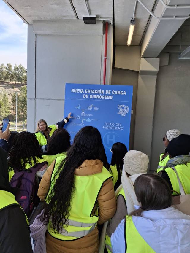 Un grupo de personas con chalecos amarillos escucha a una presentadora junto a un panel sobre la nueva estación de carga de hidrógeno.