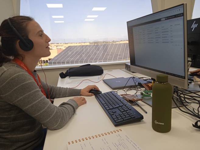 Una mujer usa auriculares y trabaja en una computadora mientras observa hacia la pantalla en una oficina iluminada.