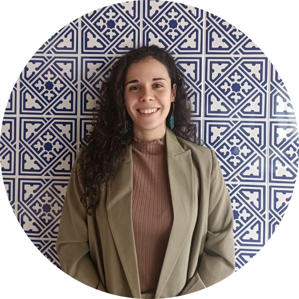 Una mujer sonriente con cabello rizado oscuro y chaqueta verde frente a un fondo de azulejos azules y blancos.