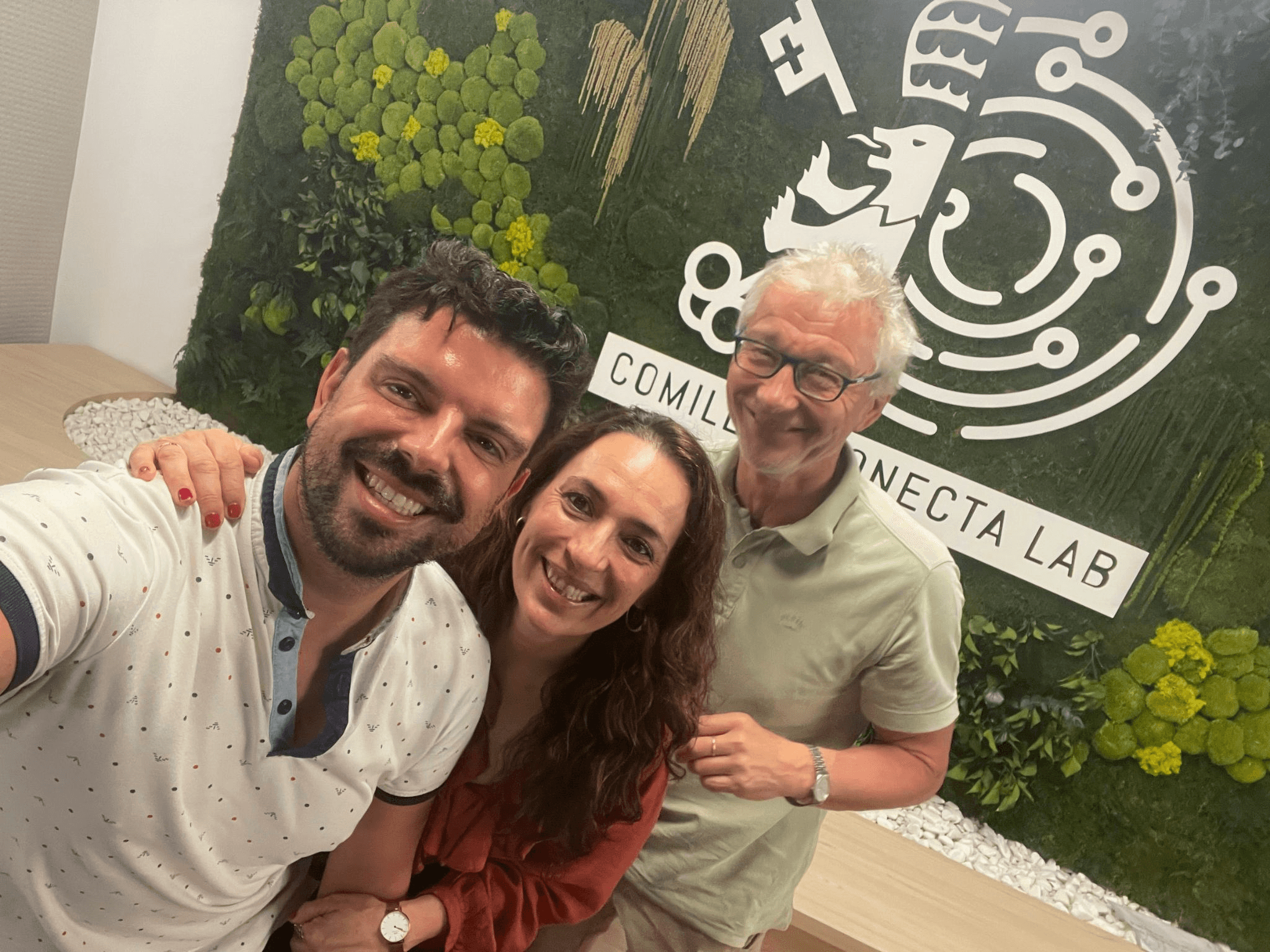 Tres personas posan juntas en un ambiente moderno y acogedor con un fondo decorado con plantas.