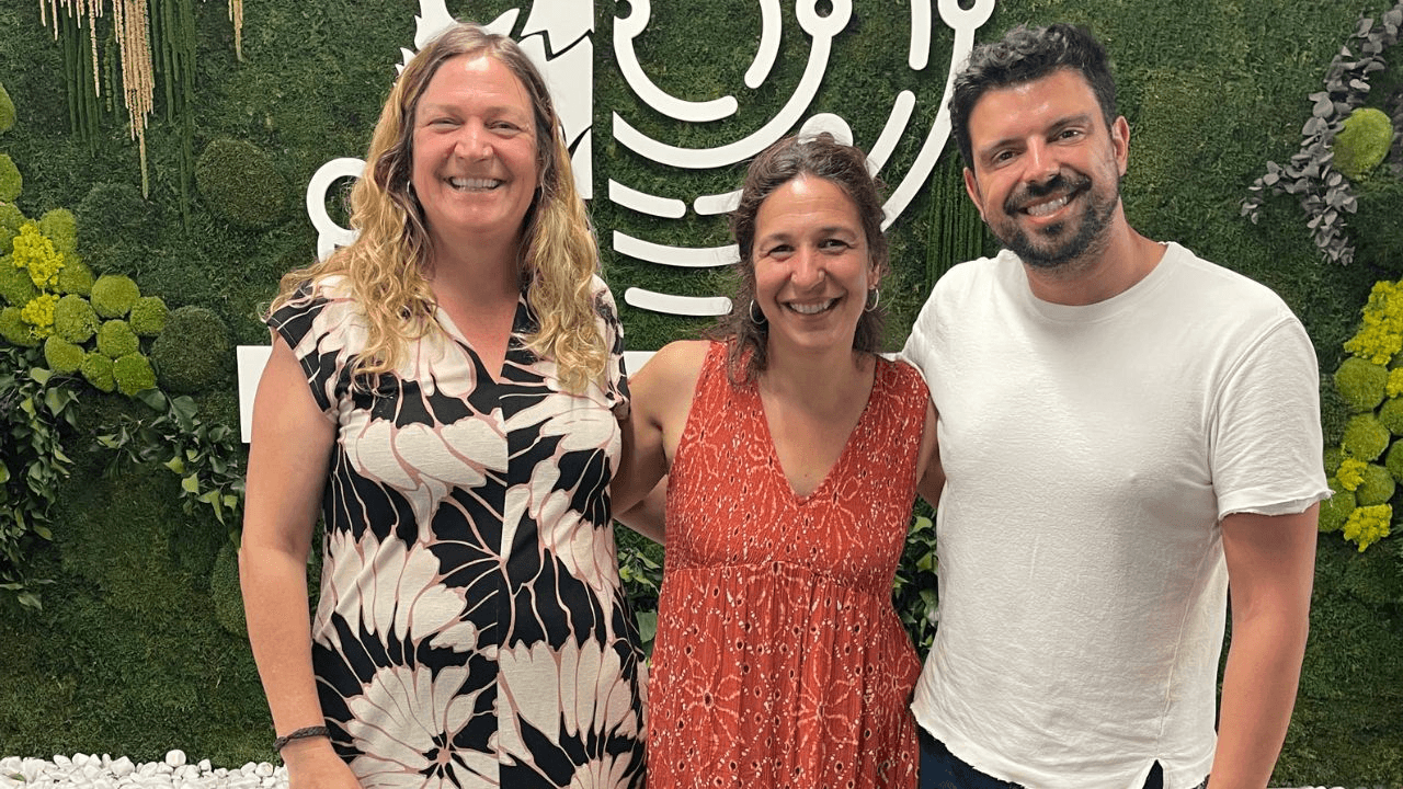 Tres personas sonrientes posan juntas frente a un fondo verde decorado con plantas.